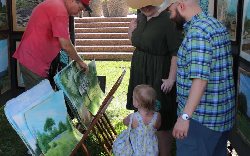 The Ricks family, right, try to pick out their favorite of several hand-painted landscape portraits during Downtown Bay City Market Day last Saturday. The painting vendor was among several braving the heat to show off their wares. Sentinel Photo/ Jessica Shepard
