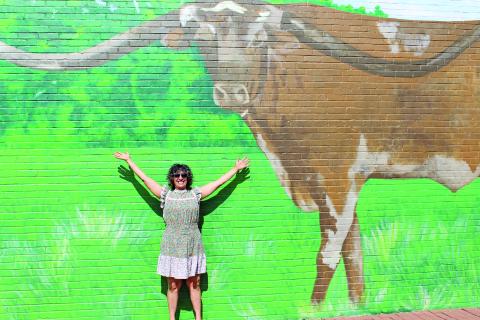 Sentinel Photo/Jessica Shepard Bay City artist Zeinab Ghais poses beneath her work-in-progress mural featuring Guinness Book of Records “Longest Longhorn Steer” 2018 winner “Sato" last Friday. Ghais has been working on the mural for almost a month and is using "point of perspective painting" to achieve a realistic look to her portrayal of the steer.
